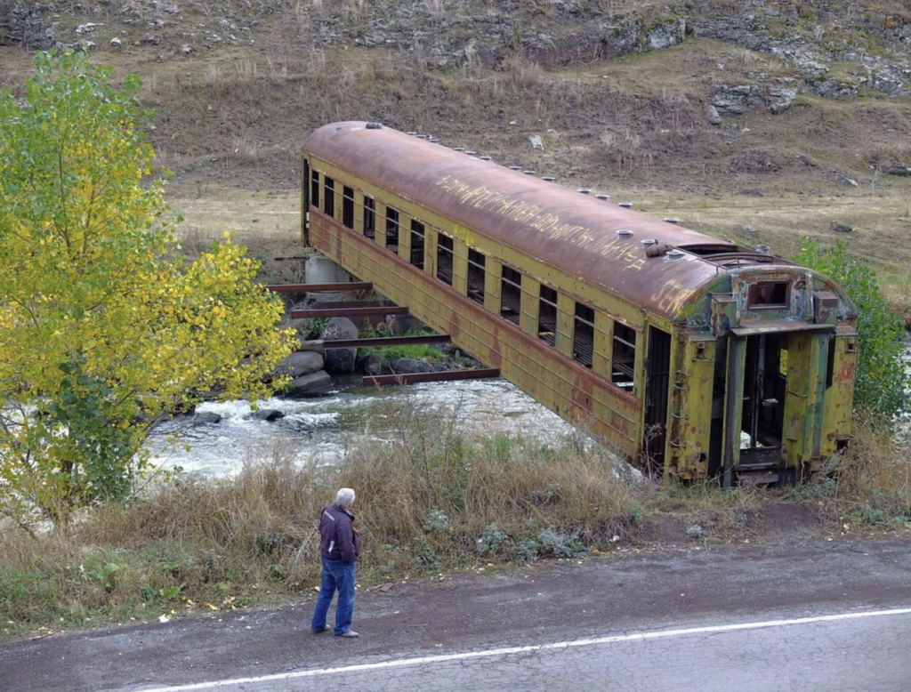 A person stands near a rusted, abandoned train car suspended over a small river by metal beams. The train car is missing its wheels and has extensive graffiti. It is surrounded by sparse vegetation and a small tree, with rocky hills in the background.