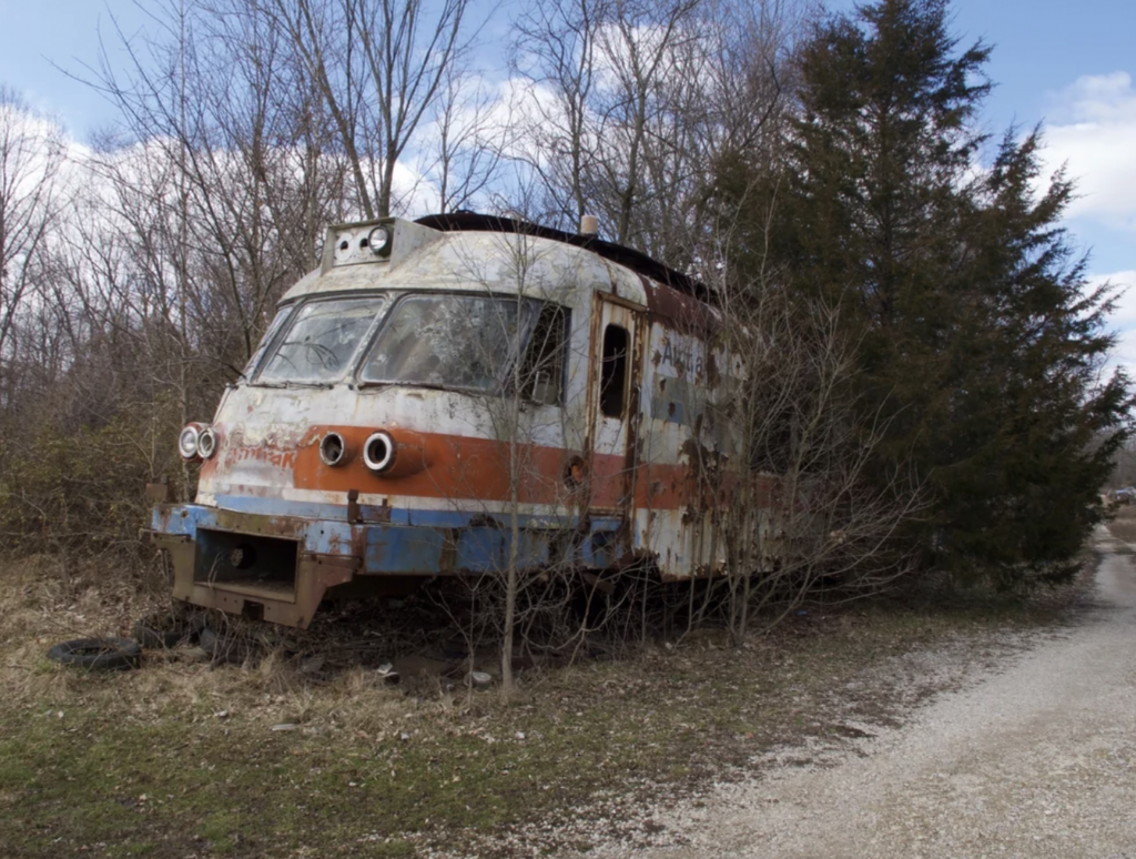 An old, rusted train locomotive abandoned in a wooded area. The metal is worn, and the train is partially overgrown with trees and bushes. The paint, which appears to have once been red, white, and blue, is peeling and faded. A dirt path runs alongside the train.