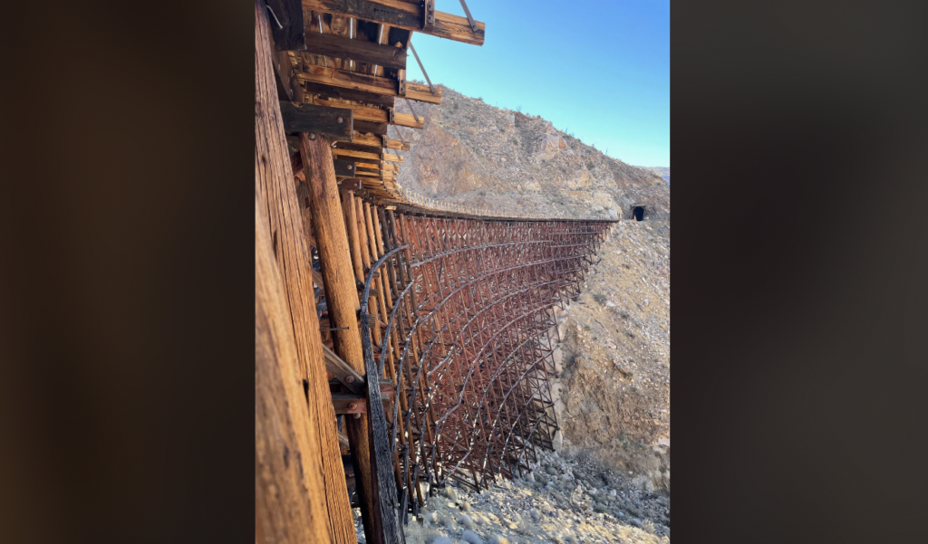 A wooden trestle bridge extends across a rocky, barren landscape under a clear blue sky. The bridge's intricate wooden beams and supports are visible, and a tunnel can be seen in the mountain on the far side of the bridge.