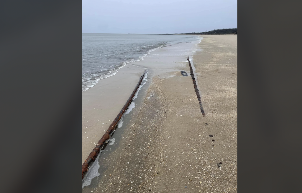 A sandy beach extends along the shoreline with gentle waves washing up against the shore. Rusted remains of a shipwreck are partially buried in the sand, creating parallel lines as they stretch towards the ocean. The sky is overcast.