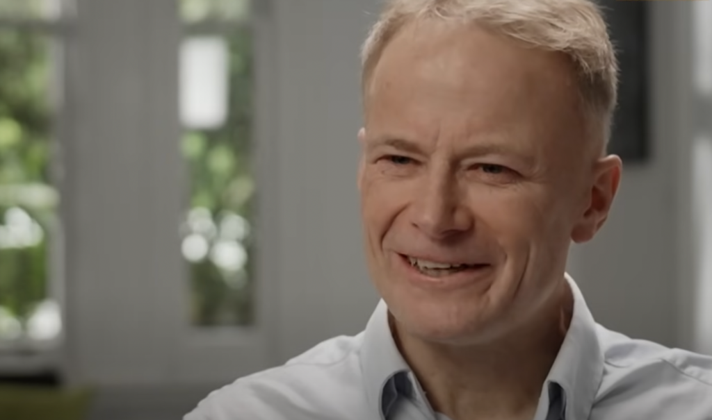 A man with short blonde hair is smiling. He is indoors, wearing a light blue shirt. The background is softly blurred but shows windows with greenery outside.