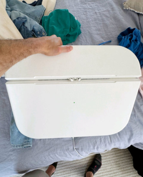 A person is holding a white device with a handle, possibly a household appliance. The device is placed on a cluttered bed with various clothes around it. The floor below shows part of a foot wearing a sandal.