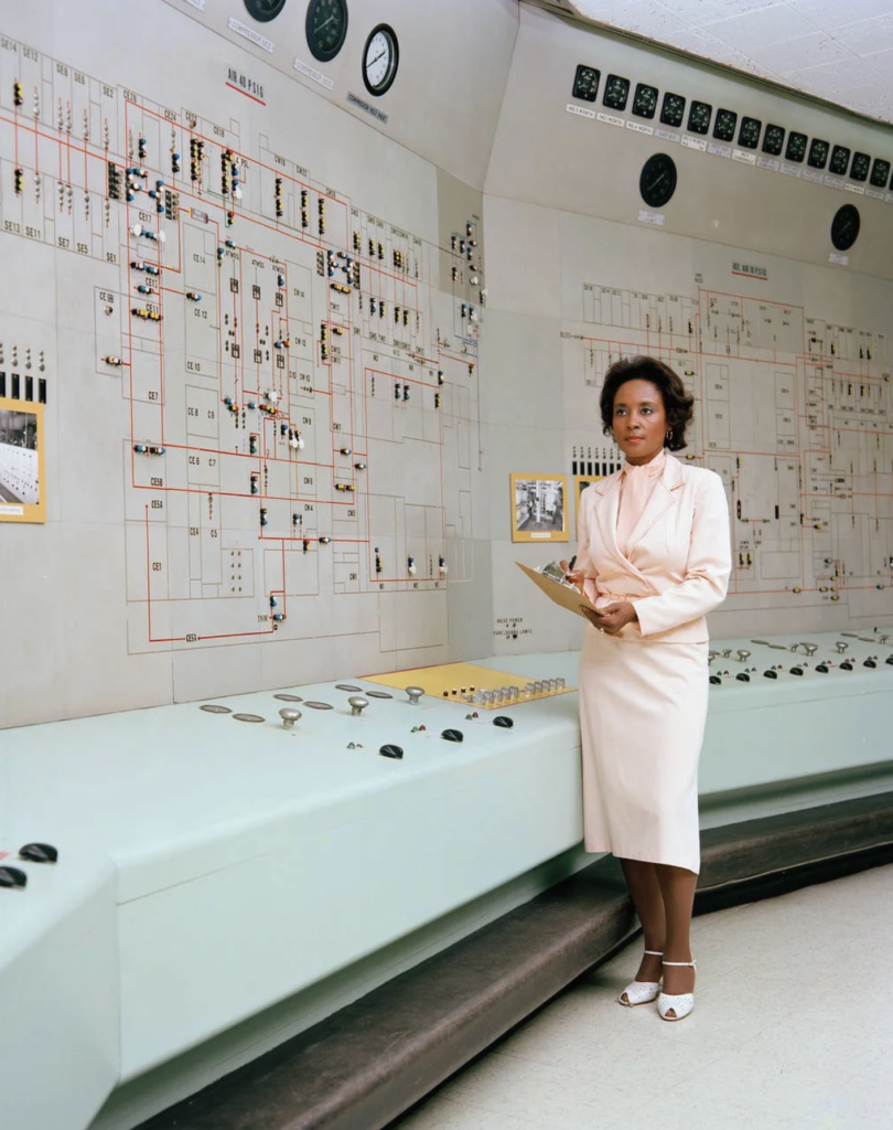 A woman in a light pink dress is standing in front of a large control panel with various buttons, switches, and dials. She is holding a clipboard and looking at the camera. The background features a detailed schematic diagram on the wall.