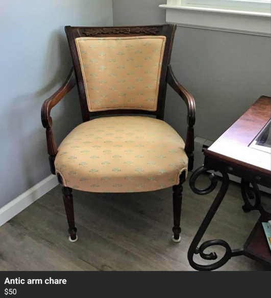 An antique wooden armchair with beige upholstery featuring a subtle patterned design. The chair has ornate carved details on the backrest and curved arms. It is positioned near a window and a small wooden table. Price listed as $50.