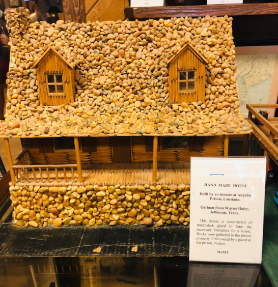 A handcrafted miniature house made of matchsticks and small pebbles is displayed. The house has two windows and a porch. A sign next to it states that it was built by an inmate at Angola Prison in Louisiana and loaned from Wayne Haley in Jefferson, Texas.