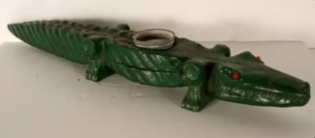 A green crocodile-shaped incense holder with red eyes stands on a flat surface. It has a circular area on its back for placing incense sticks or cones.