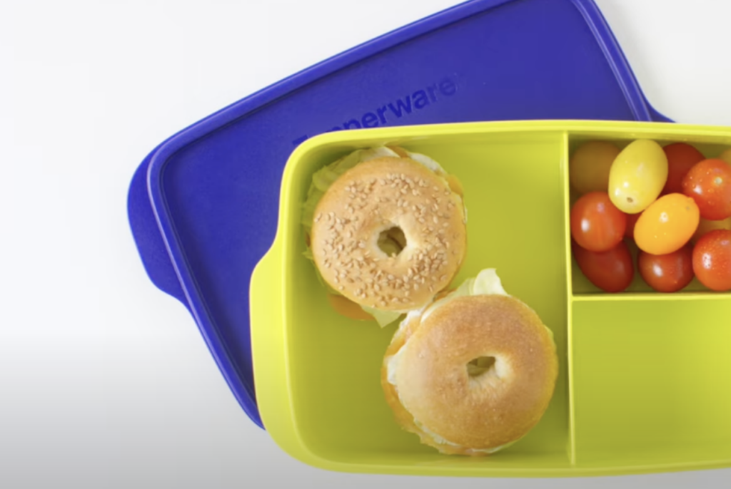 A divided Tupperware container with a blue lid, containing two sesame seed bagels with fillings on one side and a mix of red and yellow cherry tomatoes on the other side.