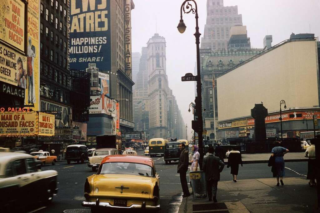 A bustling city street scene from the mid-20th century, featuring numerous vintage cars and taxis. Large advertisements and billboards dominate the buildings. People are walking on sidewalks, some with umbrellas, reflecting a lively and dynamic urban environment.