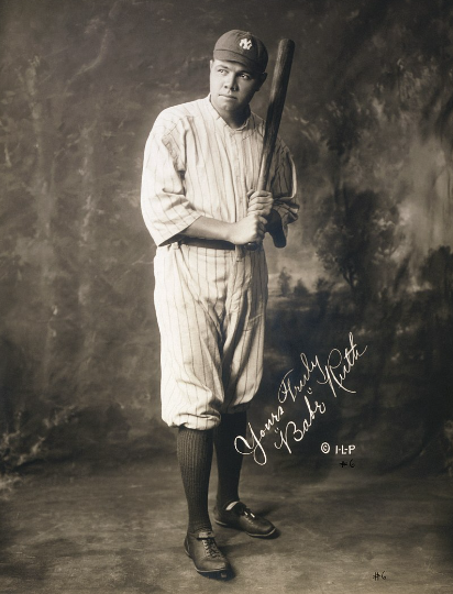 Sepia-toned photo of a baseball player wearing a classic striped uniform and cap with a bat poised on his shoulder. His uniform features long socks and cleats. The photo includes a handwritten signature, "George Herman Babe Ruth.