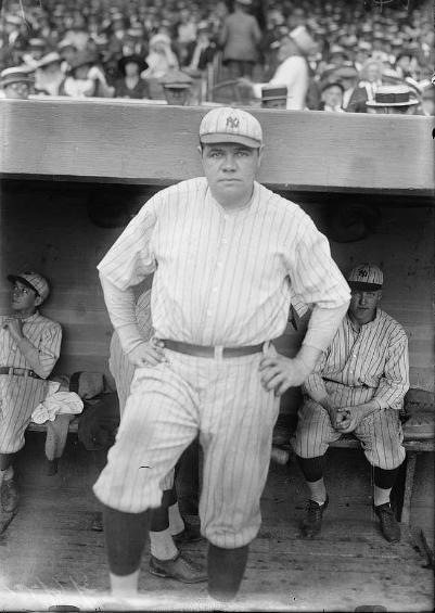 A baseball player stands in front of the dugout, wearing a striped uniform and cap. Other players can be seen sitting in the dugout behind him. A crowd fills the stands in the background. The scene appears to be from an older, historical era of baseball.