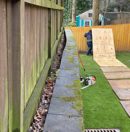 A narrow yard with a wooden fence on the left and green artificial turf on the ground. A chainsaw is placed on the turf. In the background, a person stands near a wooden ramp that leads up to a fence. Trees and a blue shed are visible.