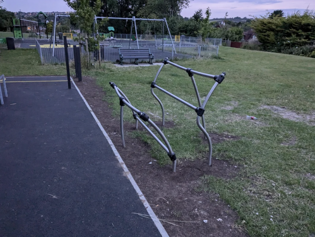 A playground featuring a modern climbing frame with metal bars. In the background, there is a set of swings, a bench, a fenced area, and trees. The sky appears to be at dusk, and the ground has both grassy and paved sections.