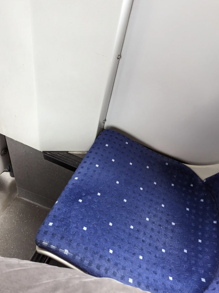 A blue fabric seat on public transportation is shown, with a pattern of small white squares. Part of a grey wall and floor is visible beside the seat. The edge of the image shows a grey object, possibly a bag or piece of clothing.