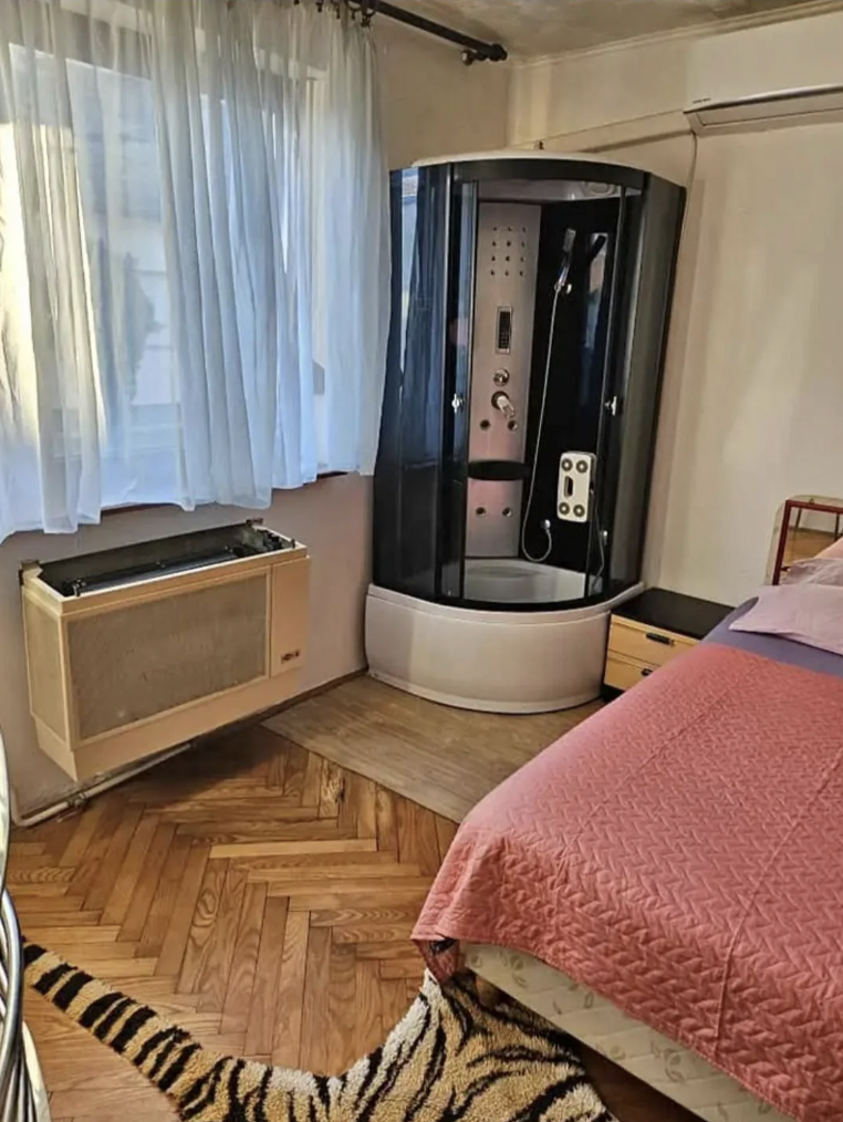 A cozy bedroom with parquet flooring features a corner shower unit with multiple settings, a pink bedspread on a wooden bed, a tiger-patterned rug, and a window with sheer white curtains. A wall-mounted air conditioning unit is situated beneath the window.