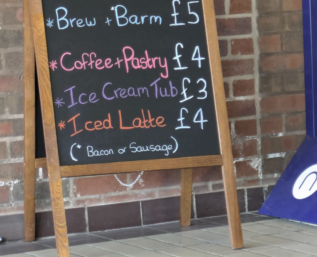 A chalkboard menu rests on a wooden stand against a brick wall. The menu lists: Brew + Barm £5, Coffee + Pastry £4, Ice Cream Tub £3, and Iced Latte £4. A footnote states: (*Bacon or Sausage). The text is written in different colors of chalk.