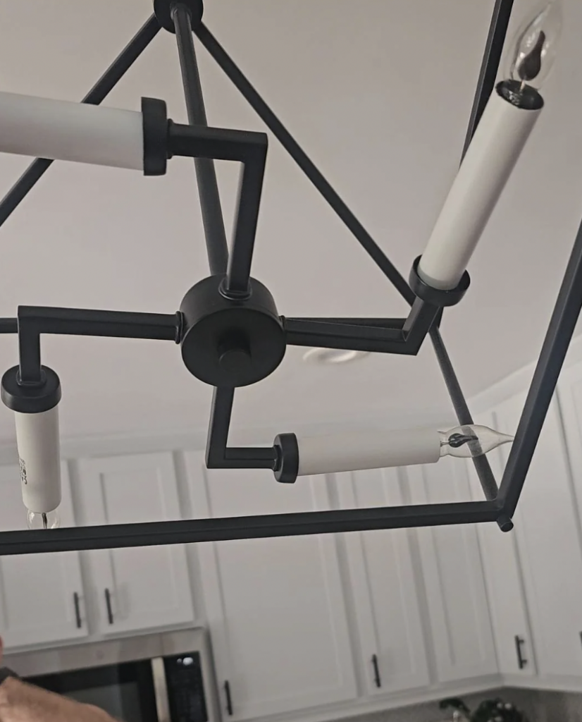 A modern, geometric chandelier with black metal framing and cylindrical white bulbs hangs from a ceiling in a kitchen. The background features white cabinetry and a glimpse of a microwave oven.