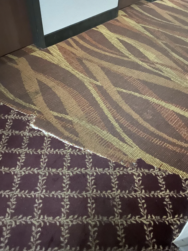 A close-up of a floor transition between two different carpet patterns. The top section features a wavy, abstract design in brown and yellow, while the bottom section shows a geometric pattern with interlocking shapes in red and white. Edges appear frayed and uneven.