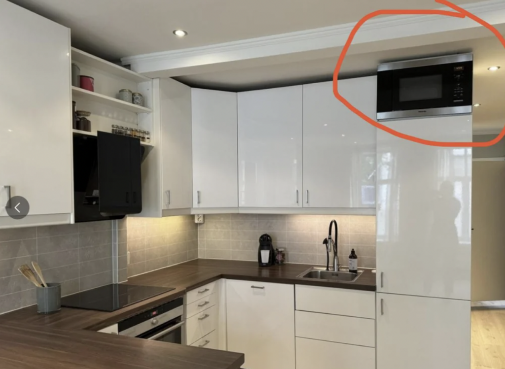 A modern kitchen with white cabinets, a tiled backsplash, and a wooden countertop. An oven and dishwasher are built into the cabinets. A microwave is circled in red and placed high above the counter. There is a sink with a faucet and soap dispenser below it.