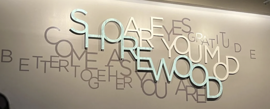 A modern wall sign features overlapping letters forming the words "SHERWOOD COME AS YOU ARE" with additional scattered phrases including "SAFETY," "GRATITUDE," "BETTER TOGETHER," "YOU MATTER," and "THRIVE." The letters are in a mix of light and darker colors.