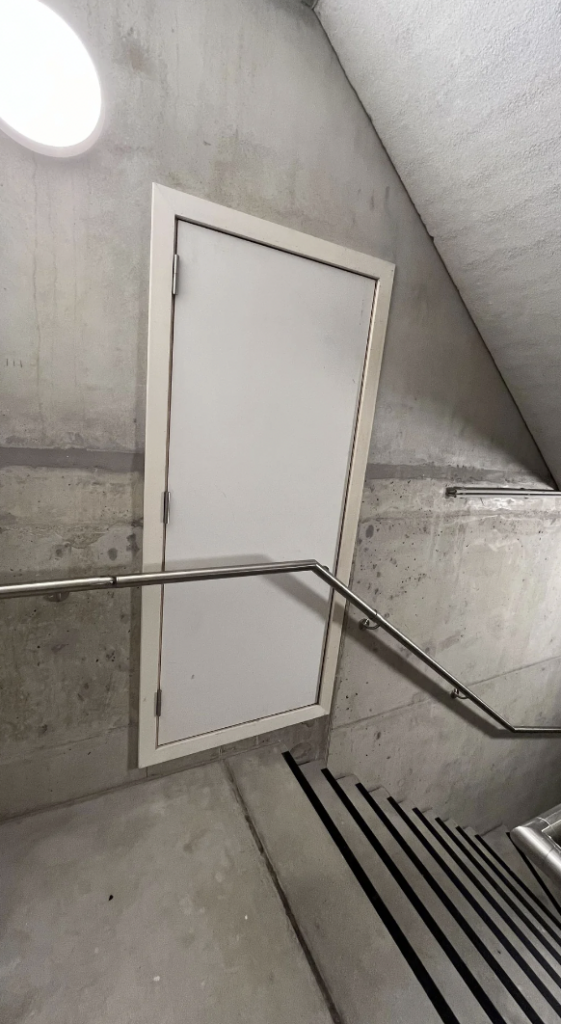 A narrow stairwell with a metal handrail on the right side and a white door at the mid-level landing. The walls and floor are made of unfinished concrete, and an overhead light illuminates the area. The stairway leads both up and down from the landing.