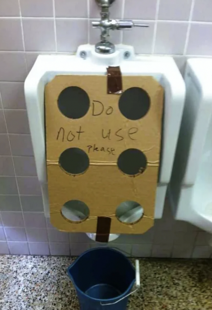 A urinal is covered with a piece of cardboard that has six large circular holes cut out. The cardboard has "Do not use please" handwritten on it. There is a strip of tape securing the cardboard, and a blue bucket is placed on the floor in front of the urinal.