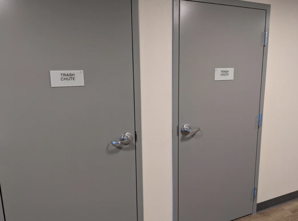 Two gray doors labeled "Trash Chute" with white signs are side by side in a beige hallway. Both doors have silver handles and are located on a wooden floor.