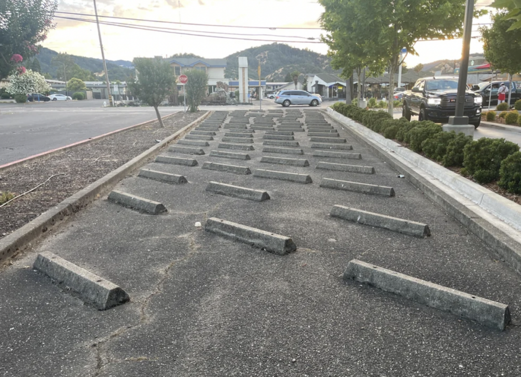 A long, narrow section of pavement with numerous concrete parking bumpers arranged in a diagonal pattern, making it unusable for parking. The area is bordered by curbs, with some greenery and trees on the sides, and vehicles parked in the background.