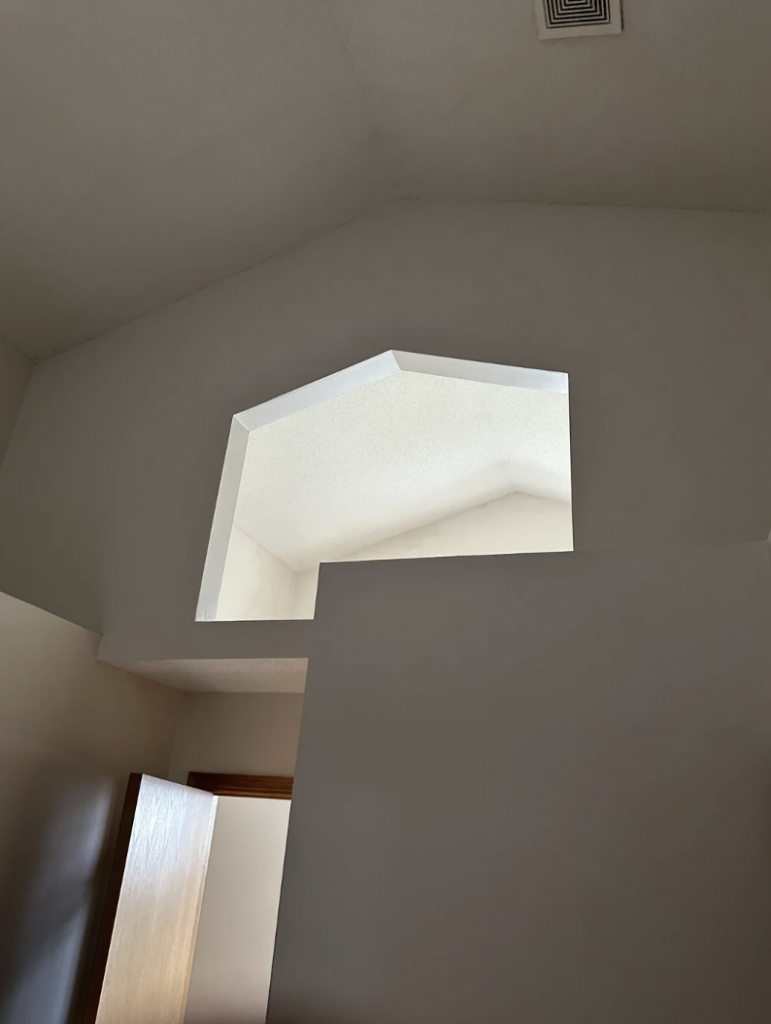 A room with high ceilings has a unique architectural feature—a large, pentagon-shaped window opening that allows natural light from an adjacent space. There’s a partially visible open wooden door and a corner of a wall in the foreground.