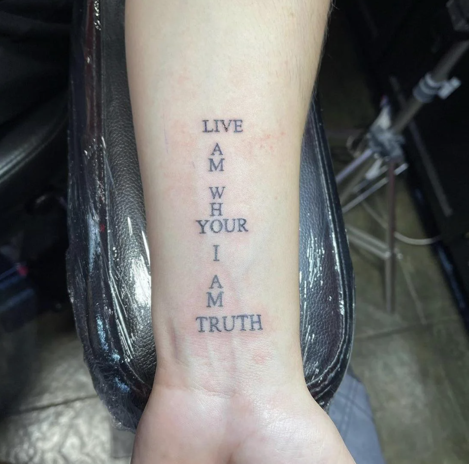 A forearm tattoo featuring vertical words: "LIVE AM WH YOUR I AM TRUTH" in bold letters. The arm rests on a black chair, and the background includes a tattoo studio with equipment partially visible.