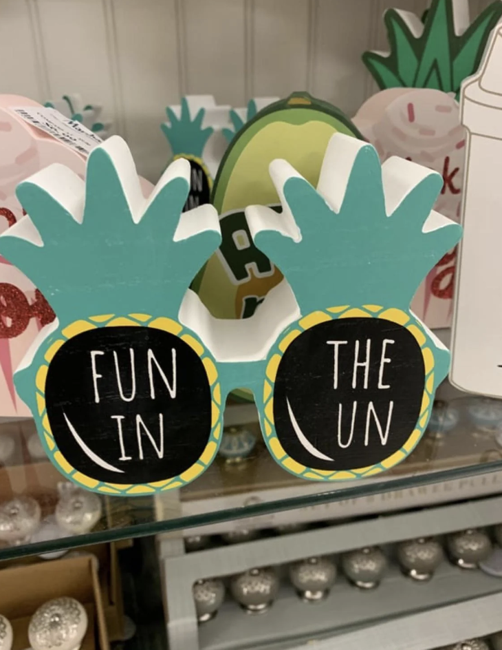 A decorative item in the shape of sunglasses with pineapple frames. The lenses have the words "FUN IN THE SUN" written on them. The background contains other tropical-themed decorations.