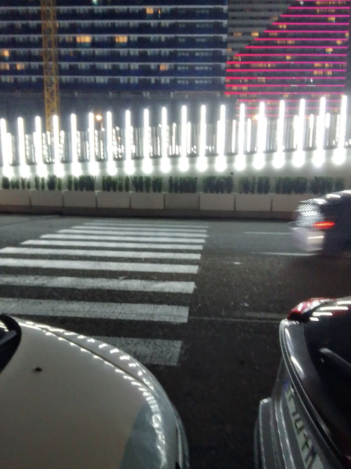 A busy road at night with a visible crosswalk. Bright vertical lights line the road, contrasting with the dark sky. Blurred headlights indicate moving cars. Modern buildings with illuminated windows and decorative light patterns are in the background.