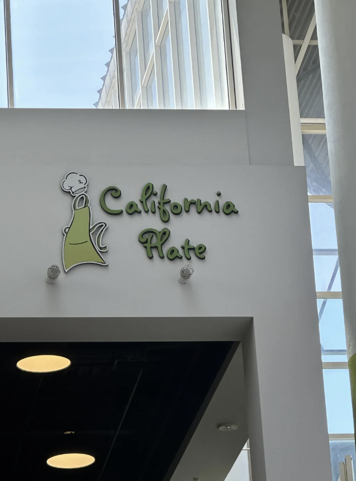 A white sign with green text reads "California Plate" beneath a graphic of a chef wearing a green apron and chef's hat. The sign is mounted above a restaurant entrance, with a high ceiling and large windows visible in the background.