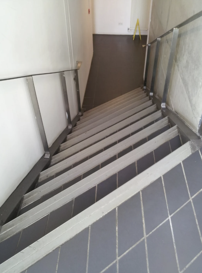 A downward view of a staircase with gray tiled steps and metallic handrails on both sides. At the base of the stairs, there is a wet floor caution sign placed on the dark gray tiled floor. The stairwell has white walls.