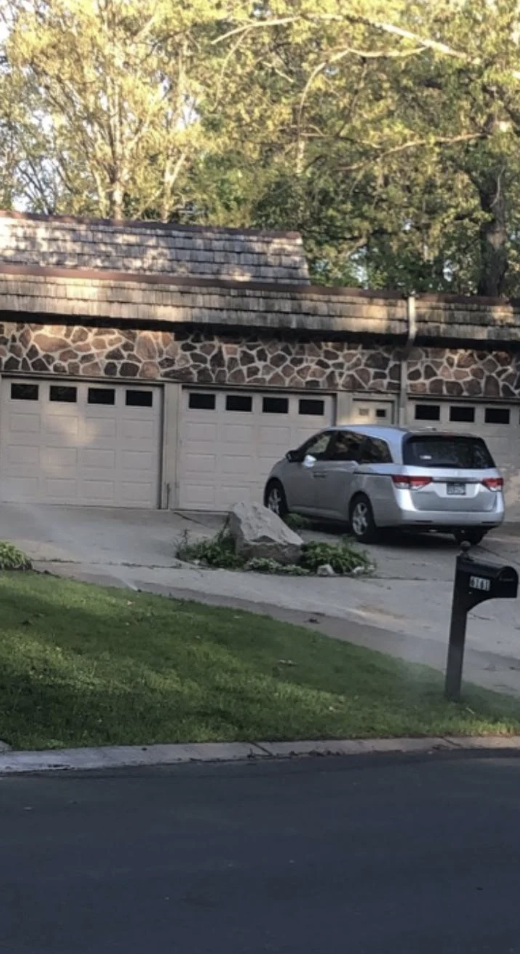 A silver minivan is parked in front of a driveway with three garage doors. The house has stone accents and a wooden shingle roof. There are trees and greenery surrounding the area. A mailbox is visible near the road in the foreground.