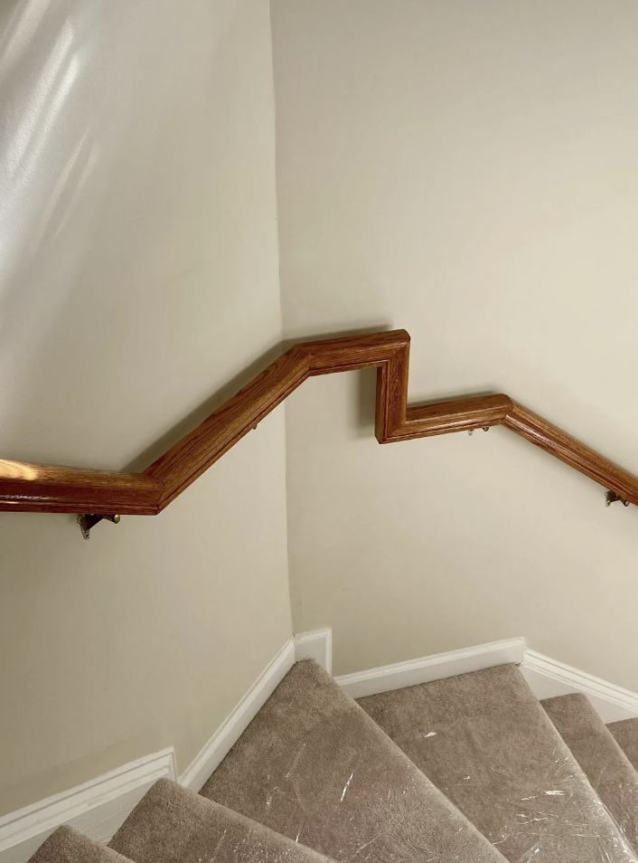 A carpeted staircase with a uniquely designed wooden handrail that zigzags down the stairs. The walls are painted a light color, enhancing the natural wood texture of the handrail.