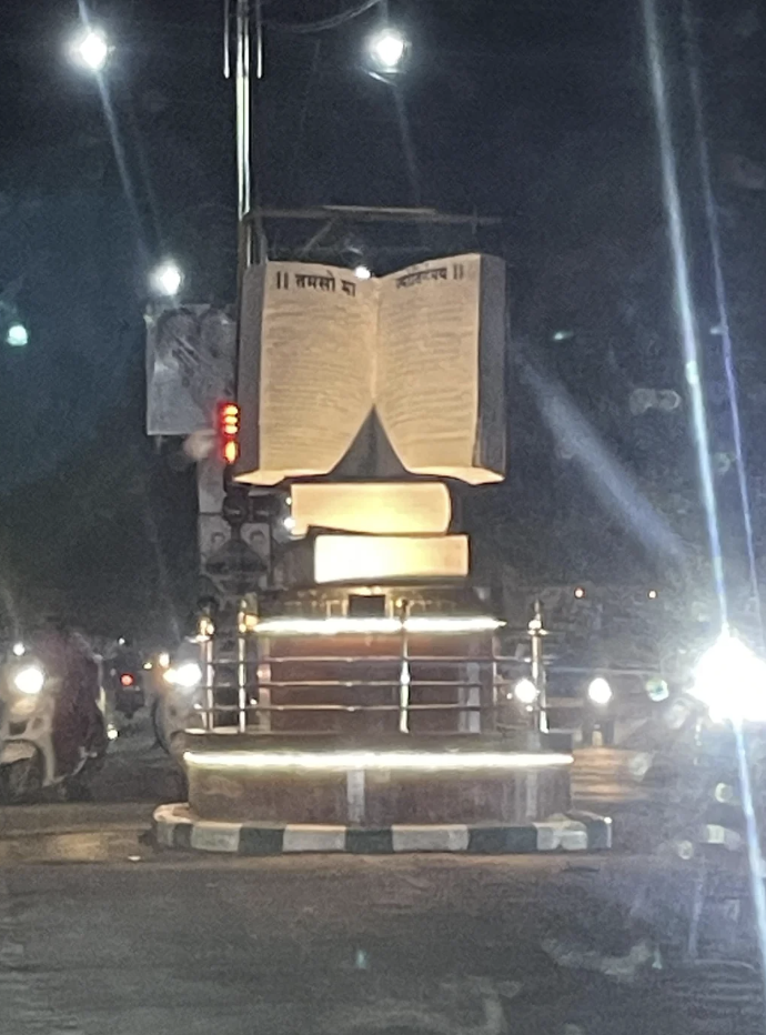 A nighttime view of a well-lit roundabout featuring a large open book sculpture mounted on a pedestal. The book is inscribed with text in an Indian script. Surrounding the roundabout are vehicles with headlights on, indicating traffic.