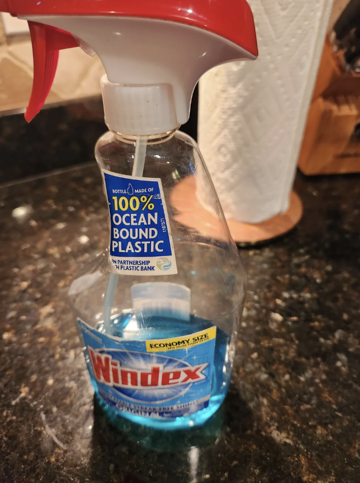 A bottle of Windex cleaner with a red spray nozzle is placed on a dark kitchen counter with a paper towel roll in the background. The label mentions that the bottle is made of 100% ocean-bound plastic in partnership with Plastic Bank.