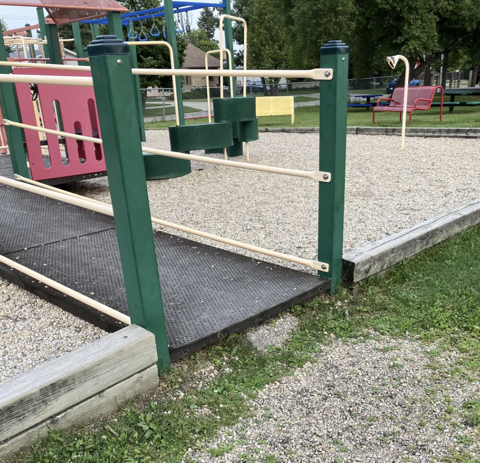 A playground entrance with a wooden ramp leading to an area filled with gravel and bordered by green posts and beige railings. In the background, play structures, benches, trees, and grass are visible.