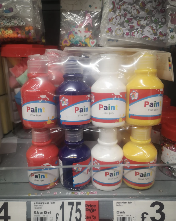 A store shelf displaying bottles of paint in four colors: red, blue, white, and yellow. The bottles are arranged in two rows, and each bottle is labeled "Paint" with a quantity of 150ml. Price tags and other small items are visible in the background.