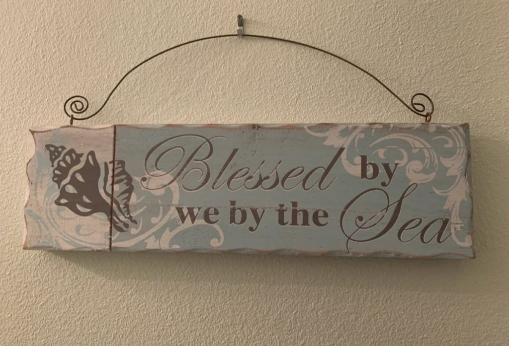 A decorative wooden sign with a rustic, sea-themed design hangs on a wall. It features the text "Blessed by we by the Sea" in elegant, script-like fonts. The sign is adorned with a seashell graphic and light blue and white colors.