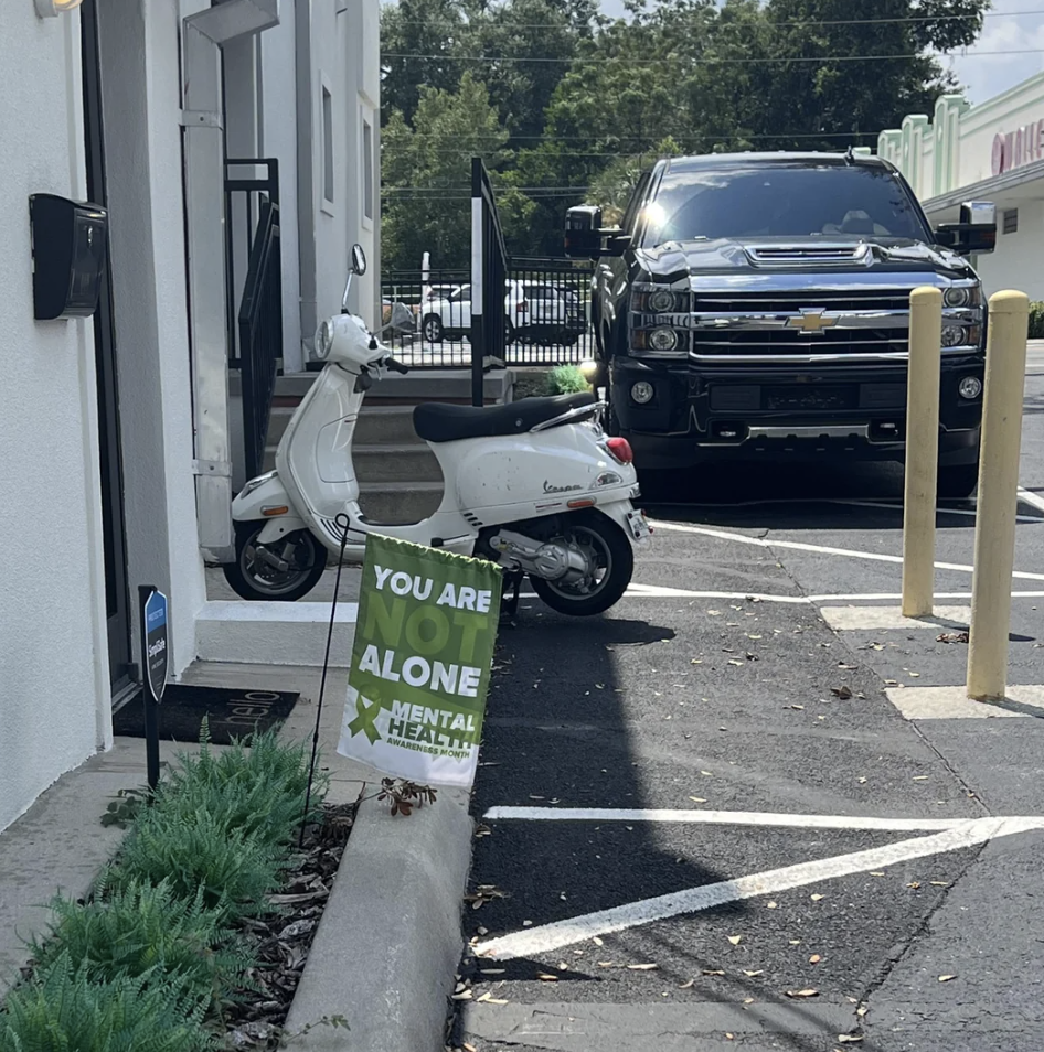 A white Vespa scooter is parked on the sidewalk next to the building entrance with a small green sign in front that reads, "YOU ARE NOT ALONE." A Chevrolet pickup truck is parked in the adjacent parking space.