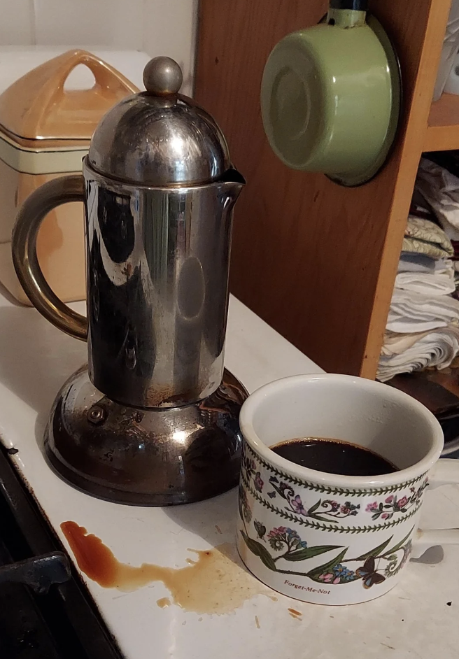 A stovetop espresso maker and a white ceramic cup with floral patterns sit on a countertop next to a wooden cabinet. The espresso maker has a metallic finish, and a coffee spill is visible on the surface below. A green mug hangs on a hook above.