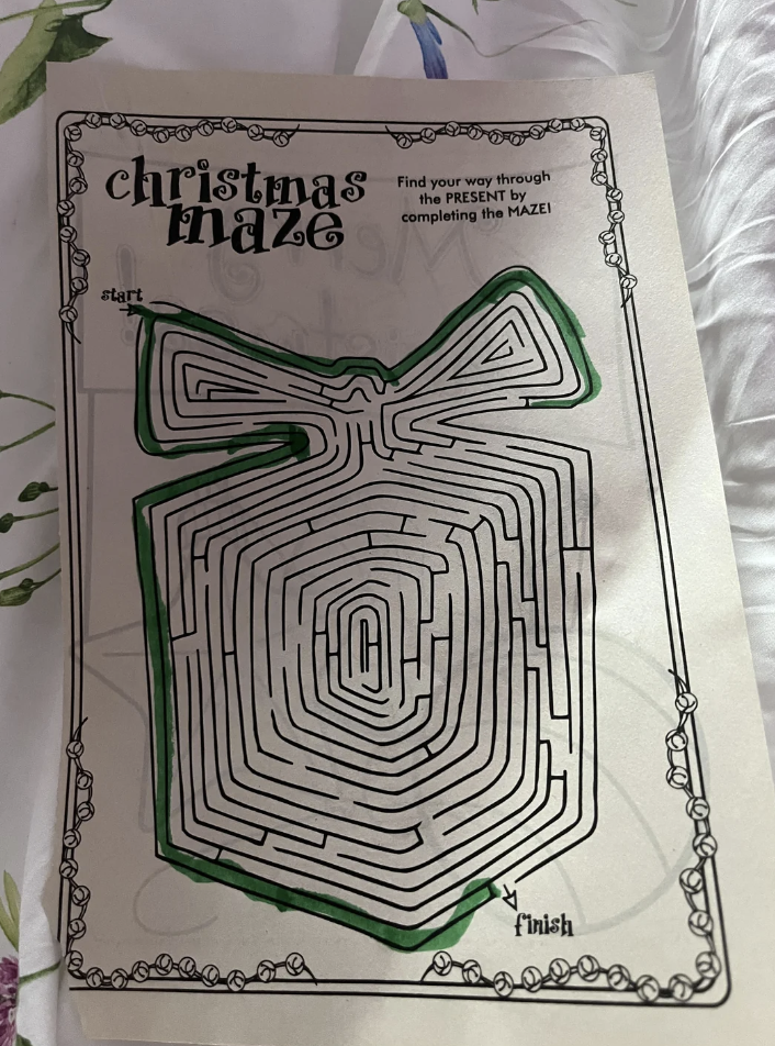 A Christmas-themed maze in the shape of a large gift with a bow. The maze starts at the upper left and ends at the bottom center. A green marker path traces the way from the start to the finish. Decorative, festive border surrounds the maze.