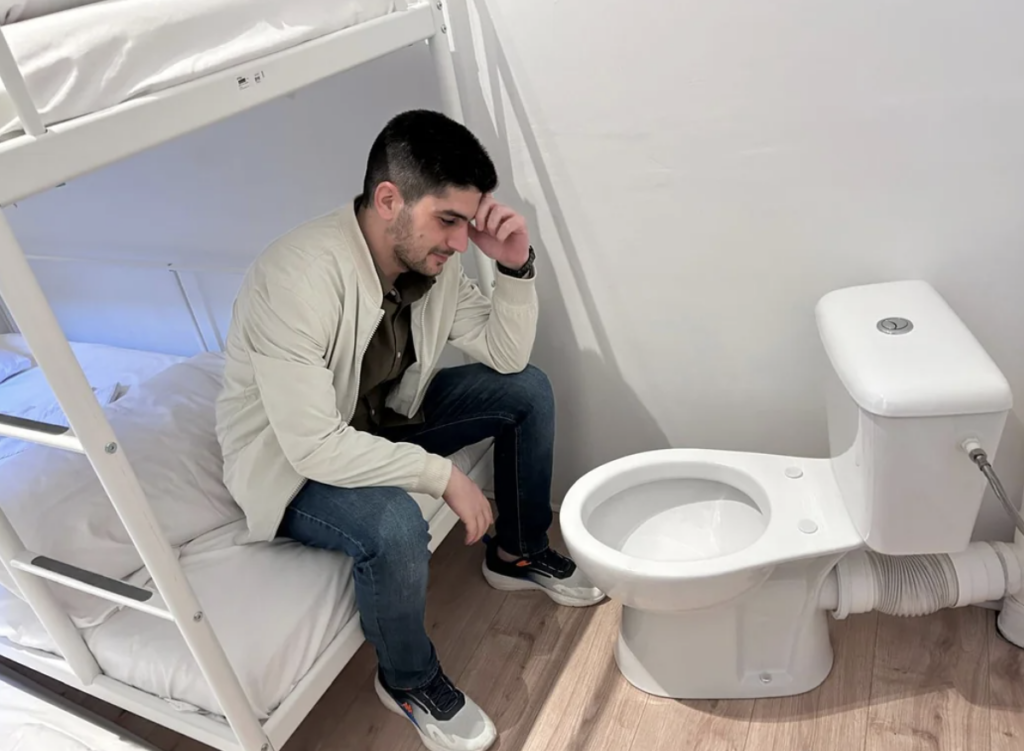 A man wearing a light jacket, jeans, and sneakers sits on a bed bunk in a small white room, holding his forehead with one hand, looking down at a toilet positioned unusually close to the bunk bed. He appears to be contemplative or concerned.