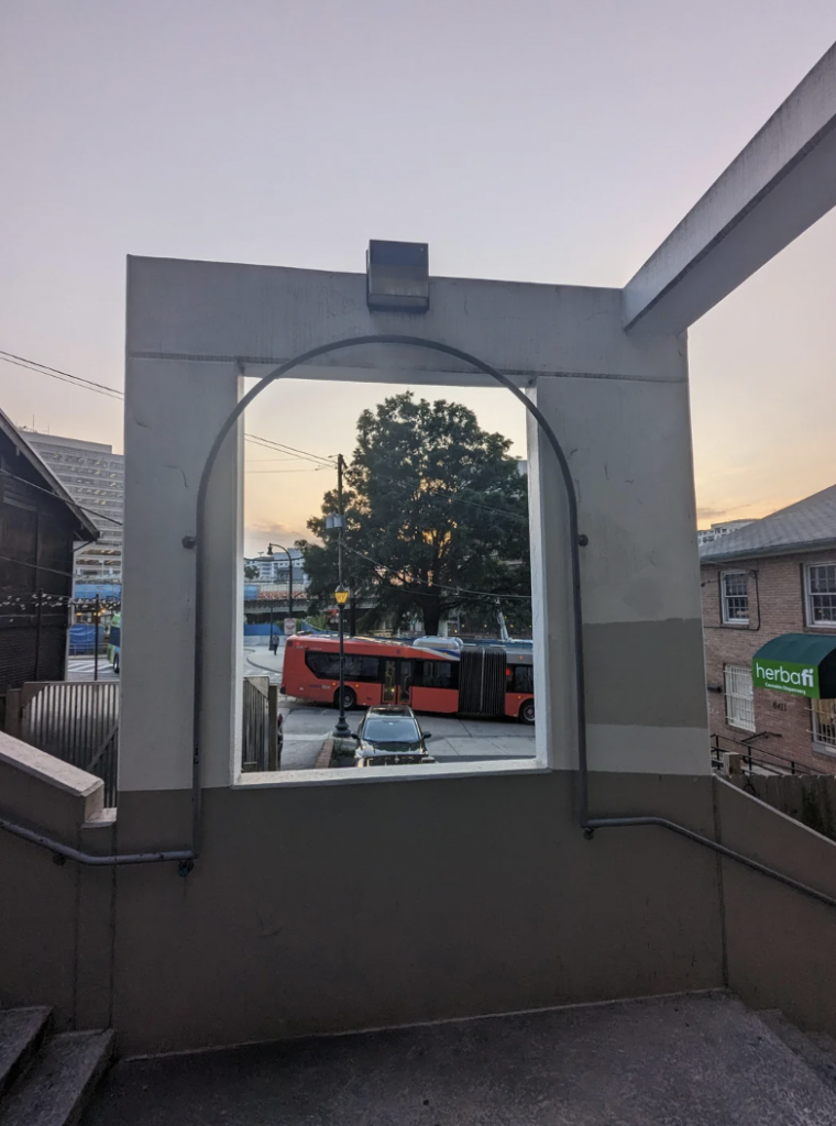 The image shows a view through a large rectangular opening in a building, overlooking a street scene. A red bus and a car are visible below, with a large tree and several buildings in the background. The sky has a subdued, early evening hue.