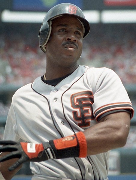 A baseball player wearing a white San Francisco Giants uniform stands at bat, holding a bat. He is wearing a helmet, black gloves with orange details, and looking intently ahead, possibly at the pitcher. The background shows a stadium with spectators.