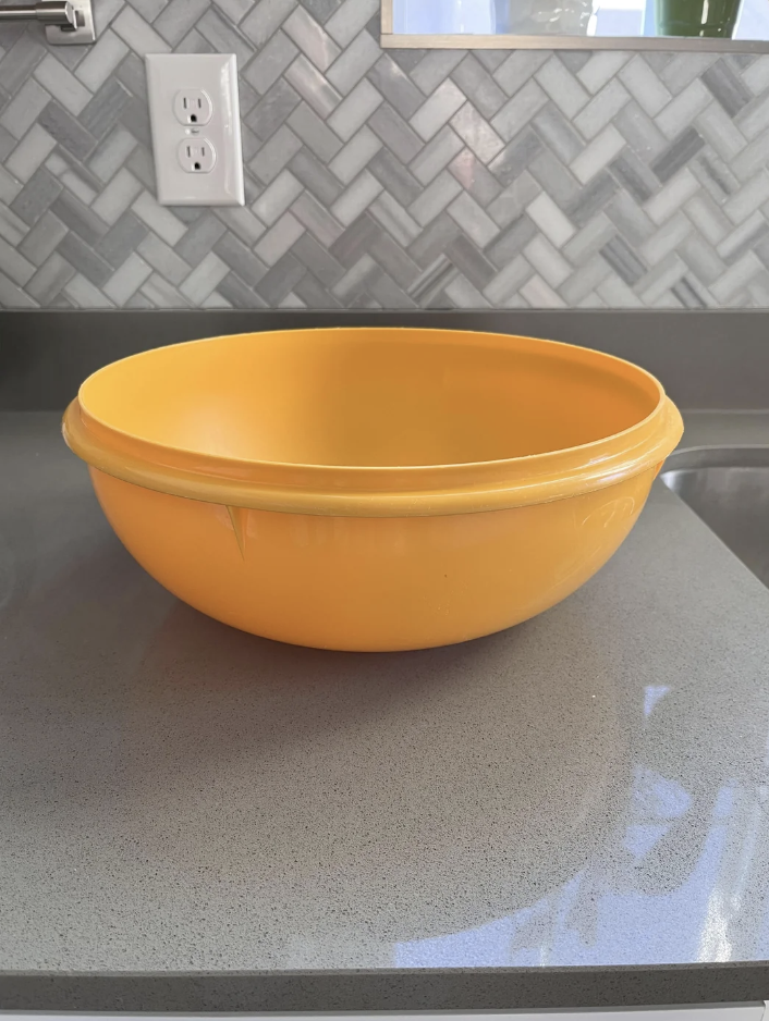 A large yellow mixing bowl sits on a gray countertop in a kitchen with a herringbone-patterned backsplash. There is an electrical outlet on the wall behind the bowl.