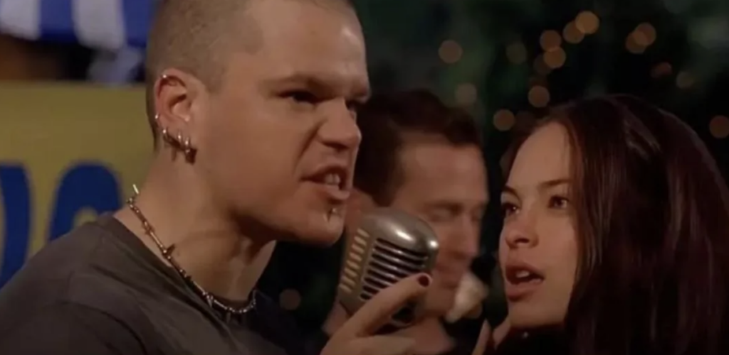 Two people are speaking or singing into an old-fashioned microphone. The person on the left has a shaved head and multiple earrings, while the person on the right has long dark hair. Both appear to be in an intense moment, possibly from a performance or a scene.