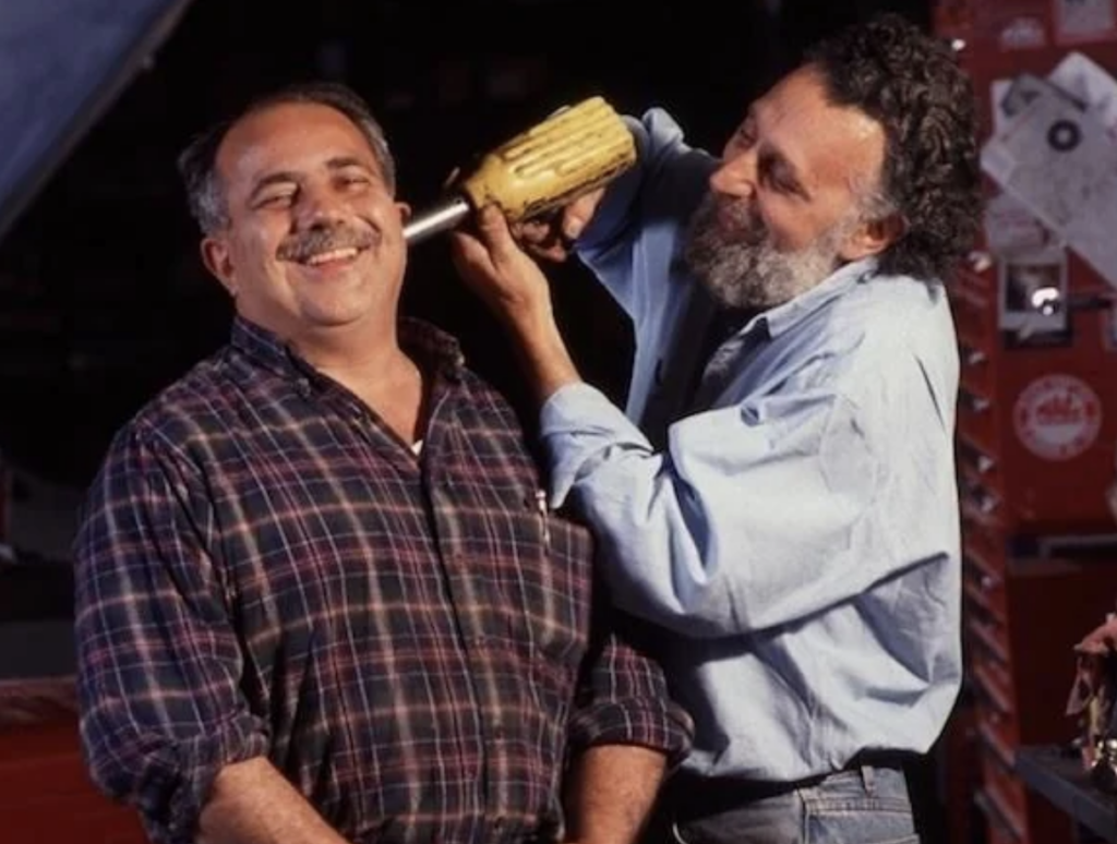 Two men in a workshop setting are sharing a playful moment. One man with a mustache is smiling while the other man, bearded, pretends to use a large power drill near his head. Various tools and red toolboxes are visible in the background.