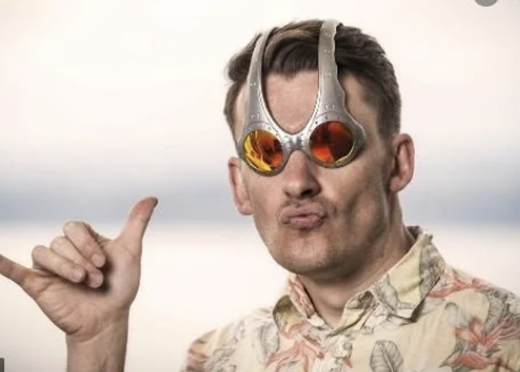 A person with short hair wearing novelty sunglasses that have exaggerated, futuristic frames and orange-tinted lenses. They are making a playful gesture with their right hand, thumb and pinky extended. They are wearing a light-colored, patterned shirt.